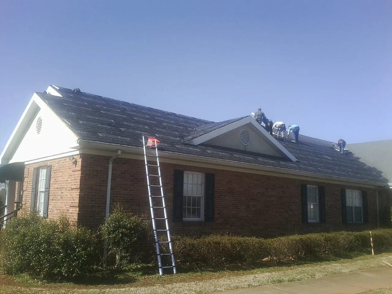 Crew tearing shingles off a brick ranch roof with a ladder set — Metal Roof Repair work by our crew in Burgoon