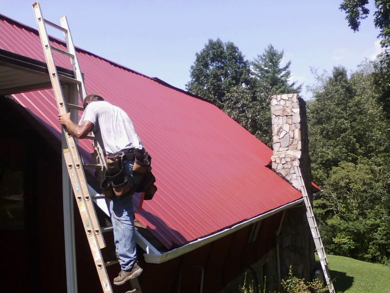 Roofer on a ladder beside a red metal roof and stone chimney — Soffit & Fascia Repair by Roofing Brothers in Burgoon